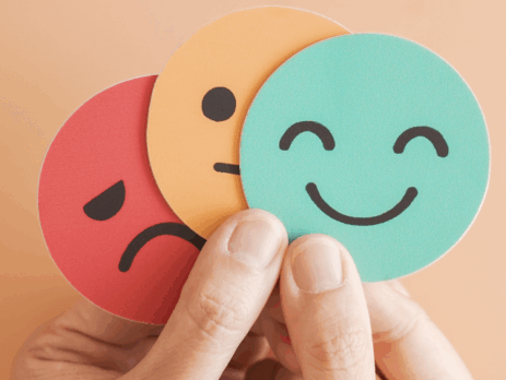 Main tenant trois pastilles rondes illustrant des émotions — un visage triste, un visage neutre et un visage souriant — sur un fond beige.