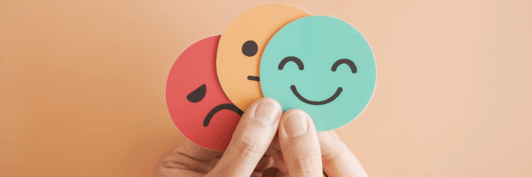 Main tenant trois pastilles rondes illustrant des émotions — un visage triste, un visage neutre et un visage souriant — sur un fond beige.