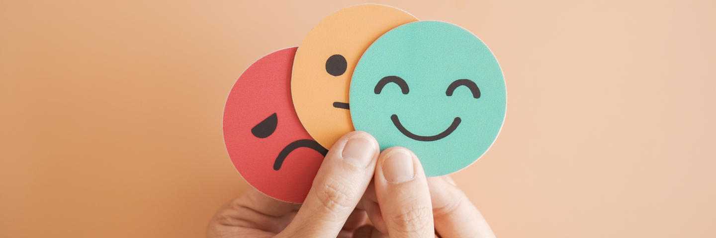Main tenant trois pastilles rondes illustrant des émotions — un visage triste, un visage neutre et un visage souriant — sur un fond beige.