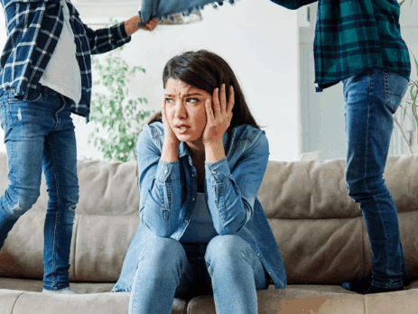 Une mère assise sur un sofa, visiblement dépassée, se tenant la tête entre les mains pendant que deux enfants sautent et crient autour d’elle dans le salon.