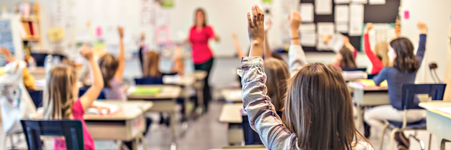 « Des élèves en classe levant la main pendant qu’une enseignante donne un cours, illustrant l’énergie et les ajustements liés au retour en classe.