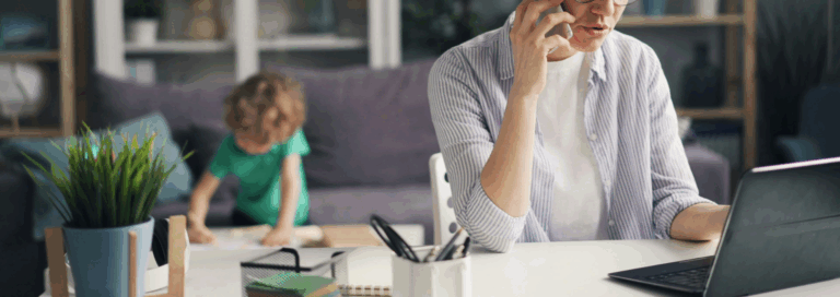 Parent en télétravail à la maison, en appel téléphonique devant un ordinateur portable, tandis qu’un jeune enfant joue ou fait une activité en arrière-plan.