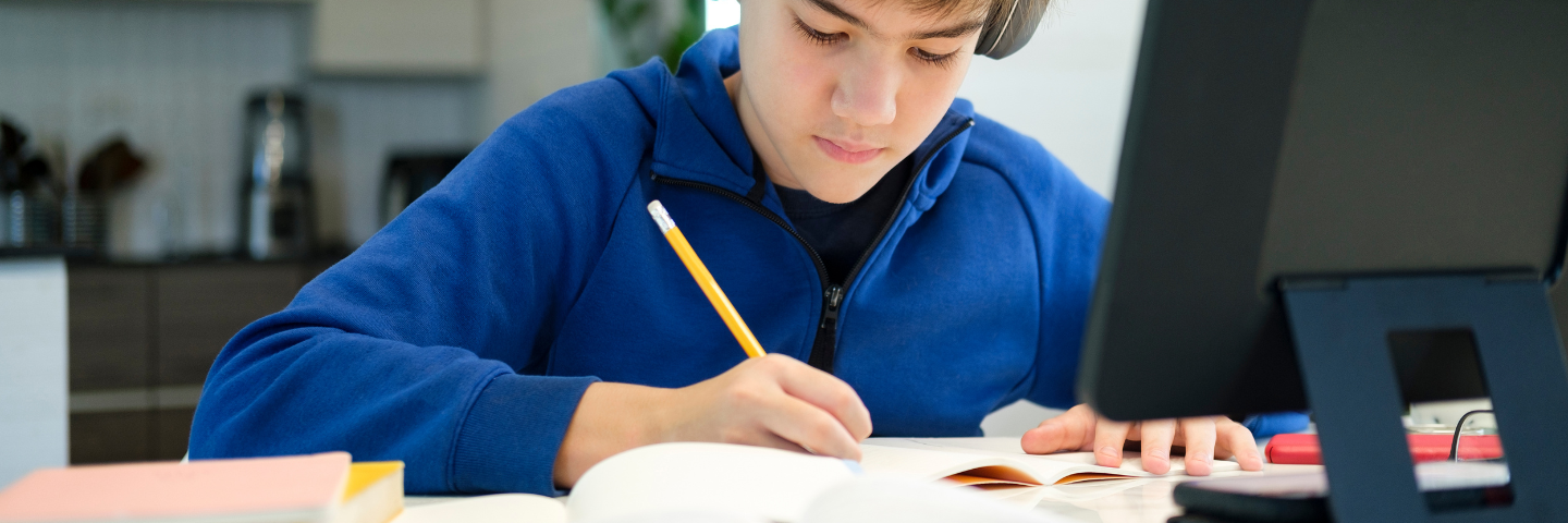 Jeune garçon concentré en train d’écrire dans un cahier à son bureau, avec un écran d’ordinateur à côté.