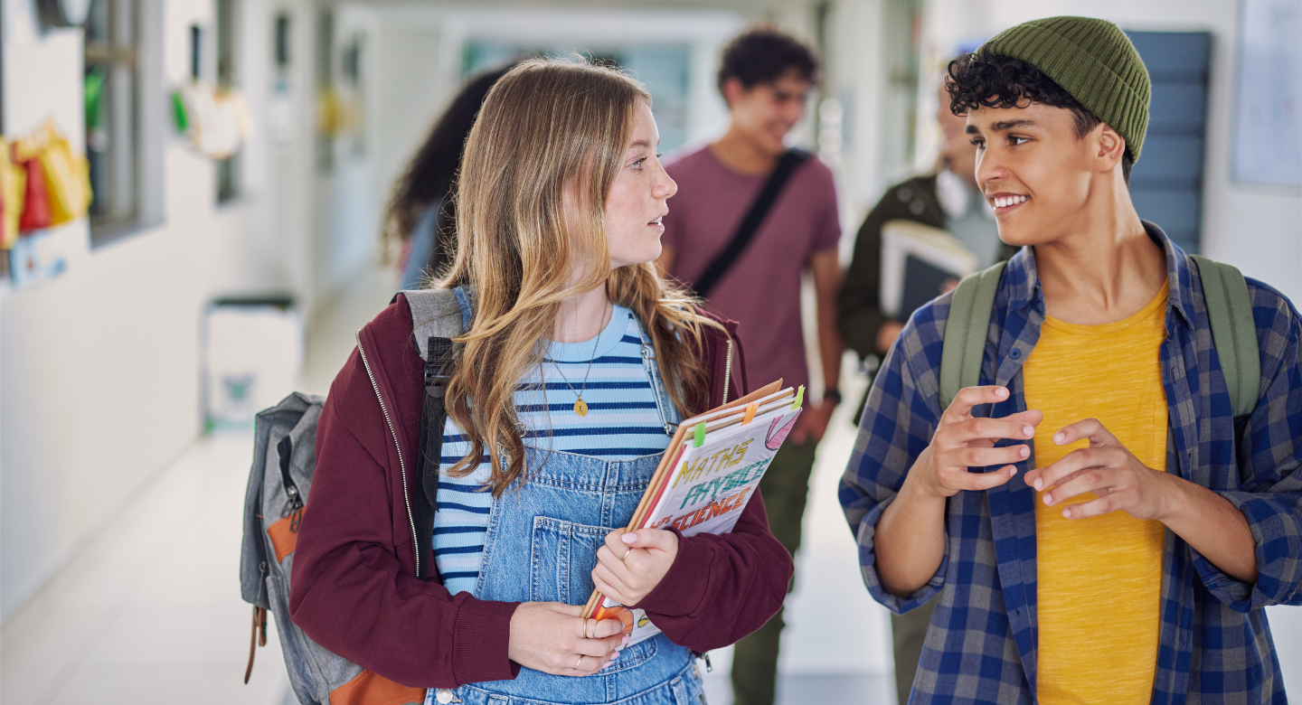 Deux élèves du secondaire discutent dans un corridor d’école, avec leurs sacs à dos et des cahiers.